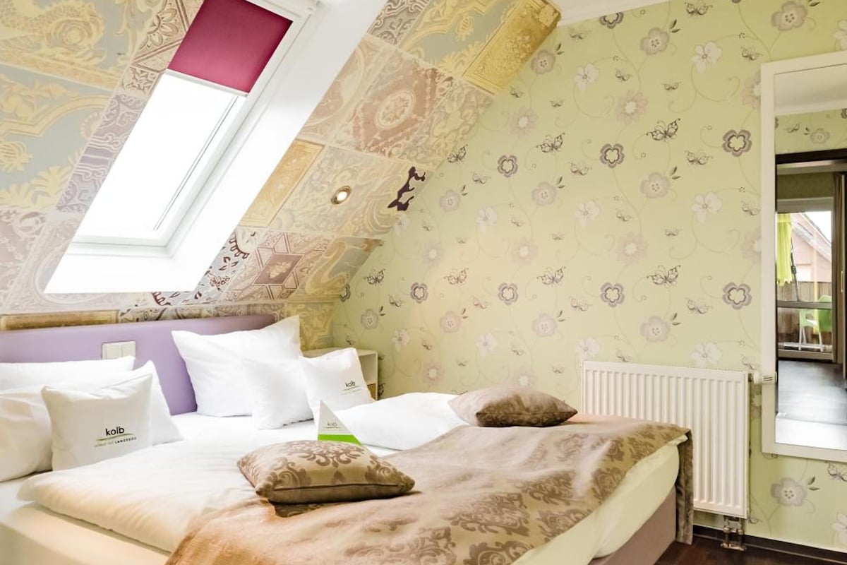 Un dormitorio con techo inclinado, tragaluz, cama con ropa blanca y estampado de papel tapiz floral.