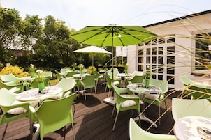 An outdoor seating area with green chairs and tables, umbrellas, and a view of trees and a building.