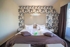 A hotel room with a large bed, two bedside tables, a wall with a pattern, and a window with curtains. - Retro Design Hotel (Langeoog)