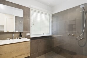 Ein modernes Badezimmer mit einem großen Spiegel, einem Waschbecken, einer Dusche mit Duschbrause und einem Fenster mit Jalousien.