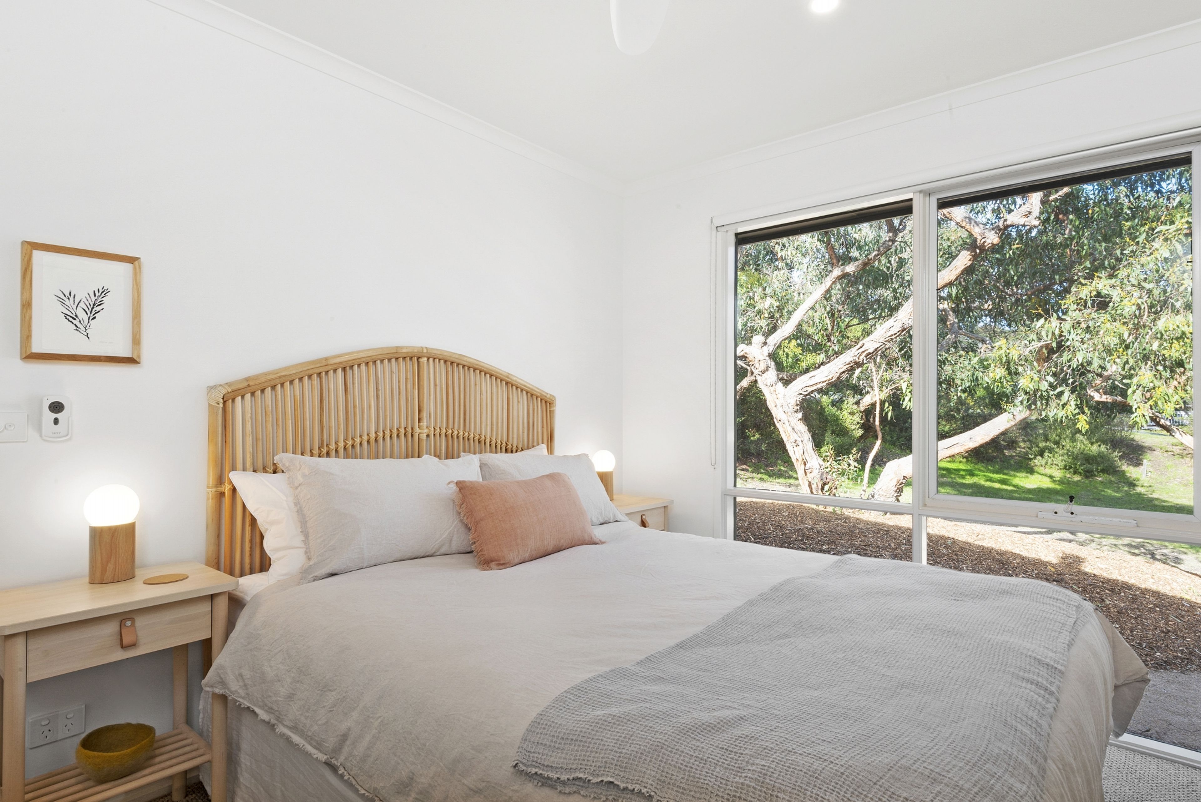 Une chambre à coucher comprenant un lit, des tables de chevet, une commode, une lampe fixée au mur et une fenêtre donnant sur des arbres.