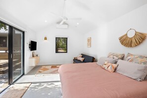 Ein modernes Schlafzimmer mit einem großen Bett, einem Deckenventilator, einem an der Wand befestigten Fernseher und Blick ins Freie durch eine Schiebetür.