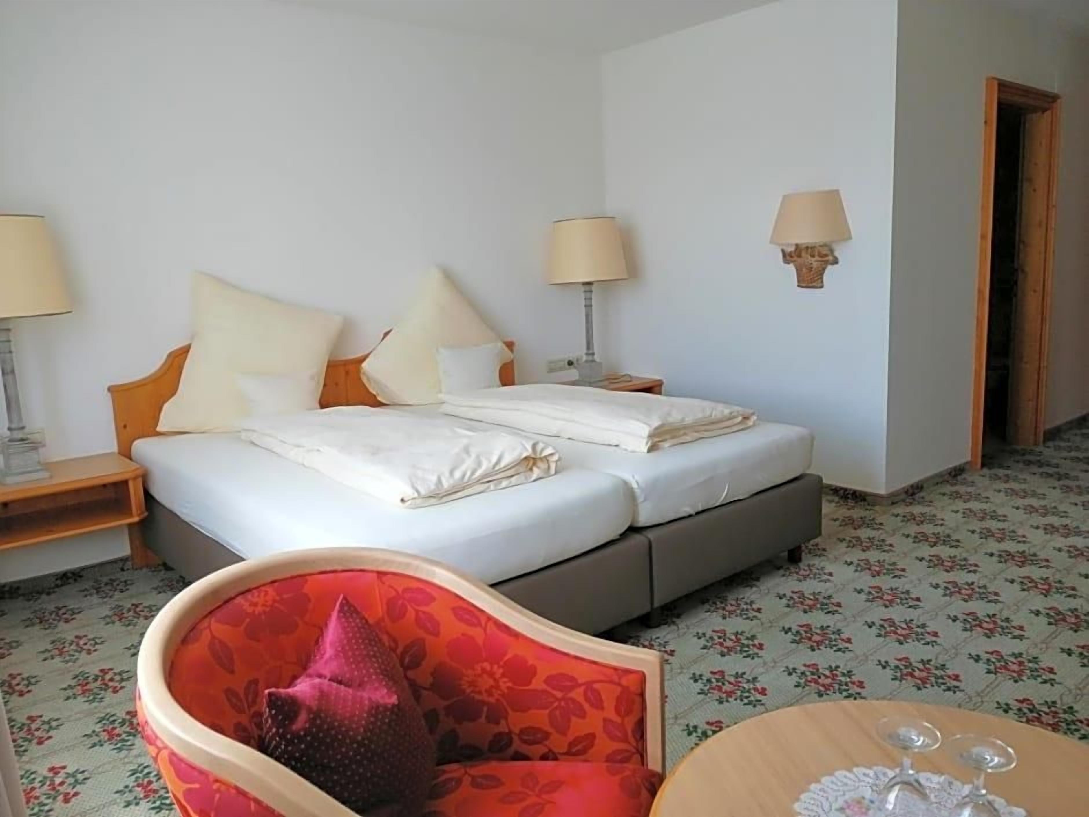 Een hotelkamer met een bed, twee bedlampen, een stoel, een tafeltje met een glas en een vloerkleed met bloemenprint.