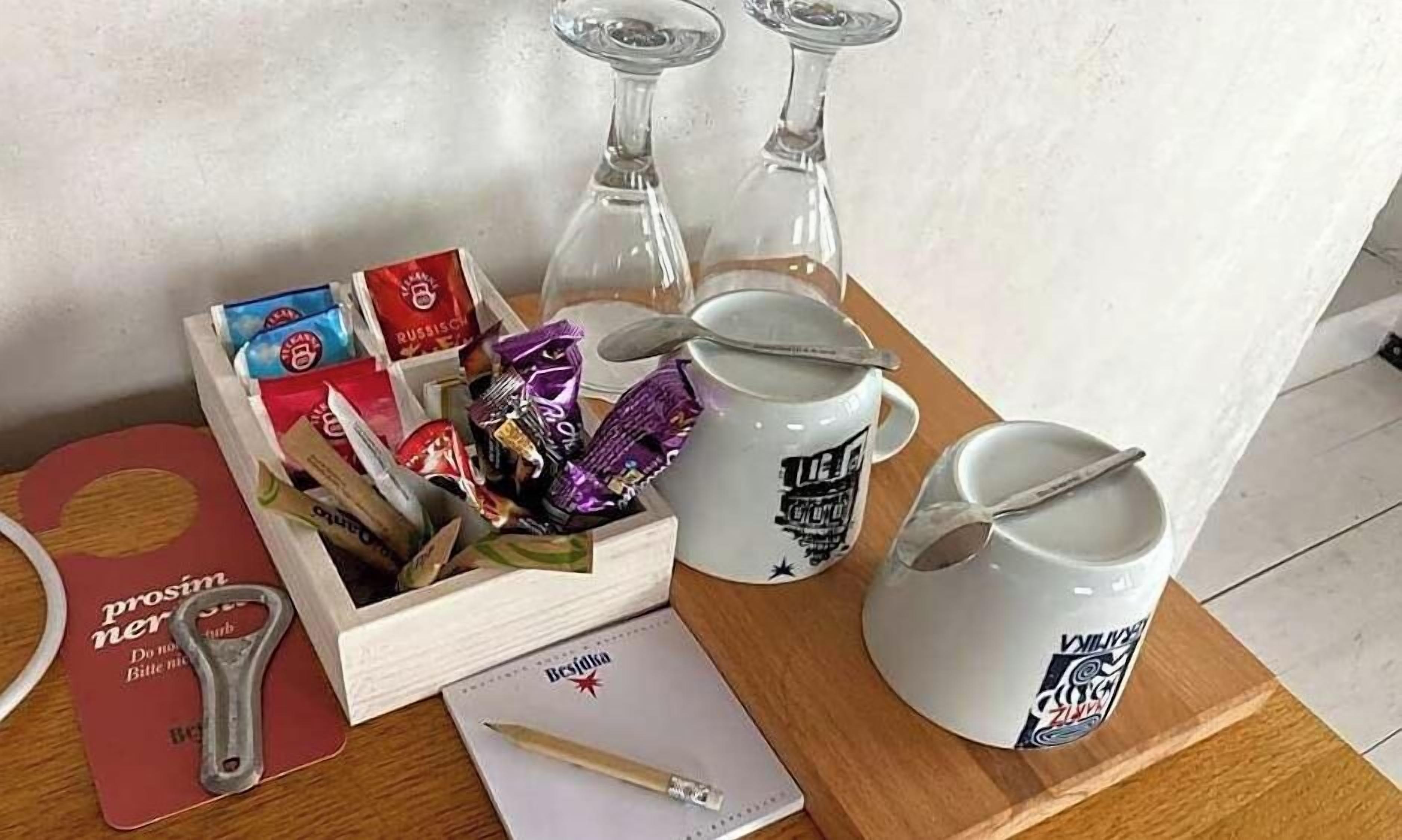 Una mesa de madera con diversos objetos, entre ellos una caja de bombones, una botella de vino, dos tazas, un sacacorchos y un bloc de notas con un bolígrafo.