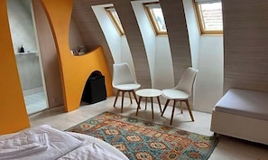 A modern bedroom with a bed, a small table with chairs, a colorful rug, and a sofa. There are skylights on the ceiling.