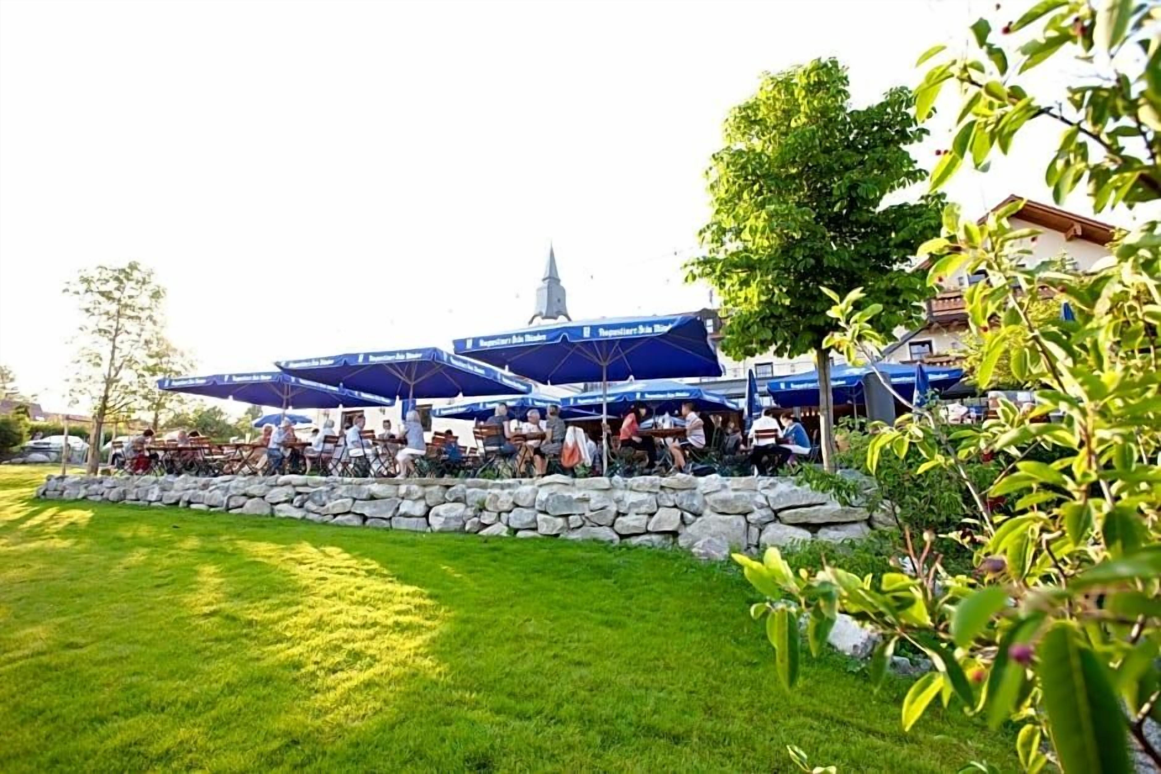Ein Sitzbereich im Freien mit Steinmauern und Sonnenschirmen, im Hintergrund ein Kirchturm und ein gepflegter Rasen.