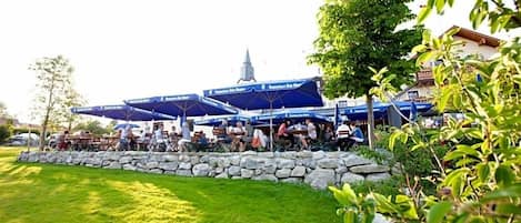 An outdoor seating area with stone walls and umbrellas, a church spire in the background, and a well-maintained lawn.