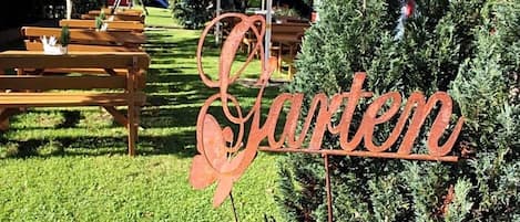 Un letrero de metal con la palabra 'Garden' en cursiva, ubicado en un área cubierta de césped con bancos de madera y un árbol pequeño.