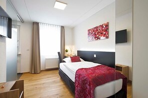 A hotel room with a bed, a desk, a chair, a window with curtains, a radiator, a wall-mounted TV, and a painting. - Landgasthof Lamm (Aalen)