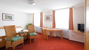 Une chambre d’hôtel comprenant un lit, deux fauteuils en osier, une petite table, un téléviseur et deux fenêtres avec des rideaux.