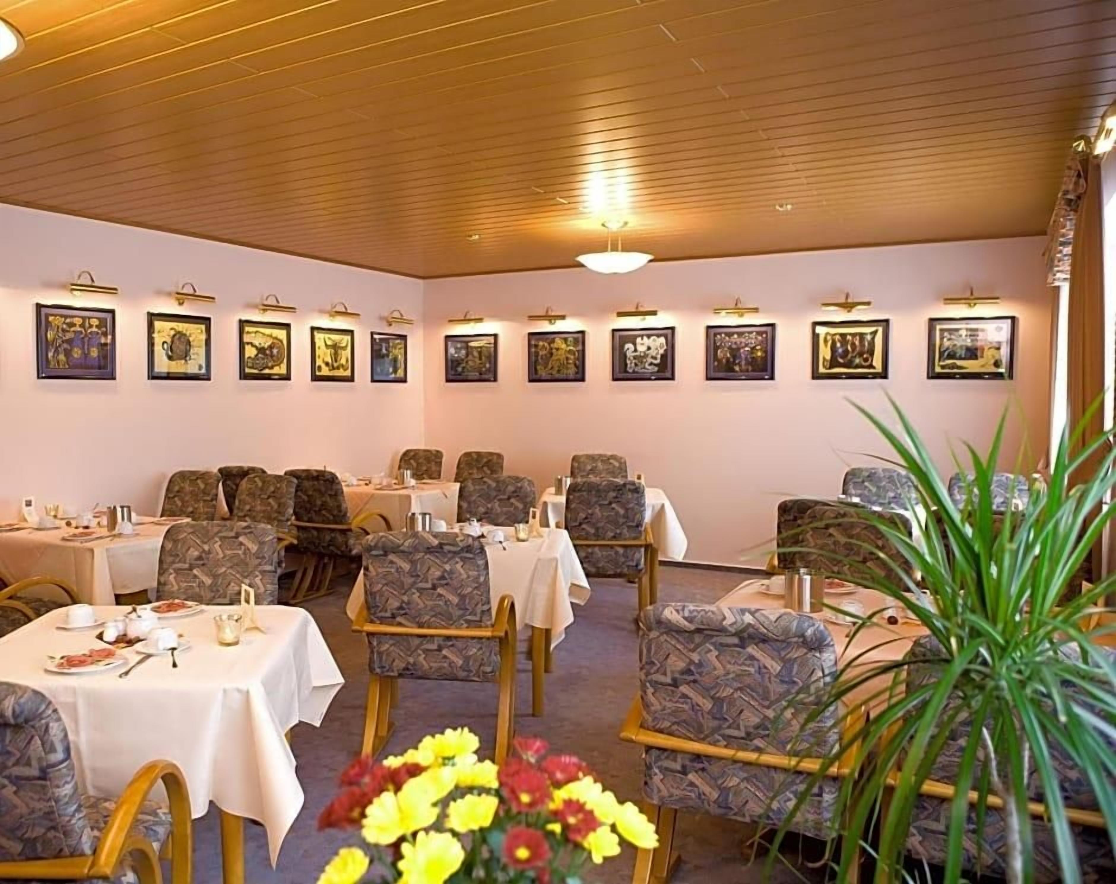 A dining area with tables and chairs, framed artwork on the walls, and a plant in the foreground.
