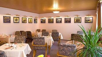A dining area with tables and chairs, framed artwork on the walls, and a plant in the foreground.