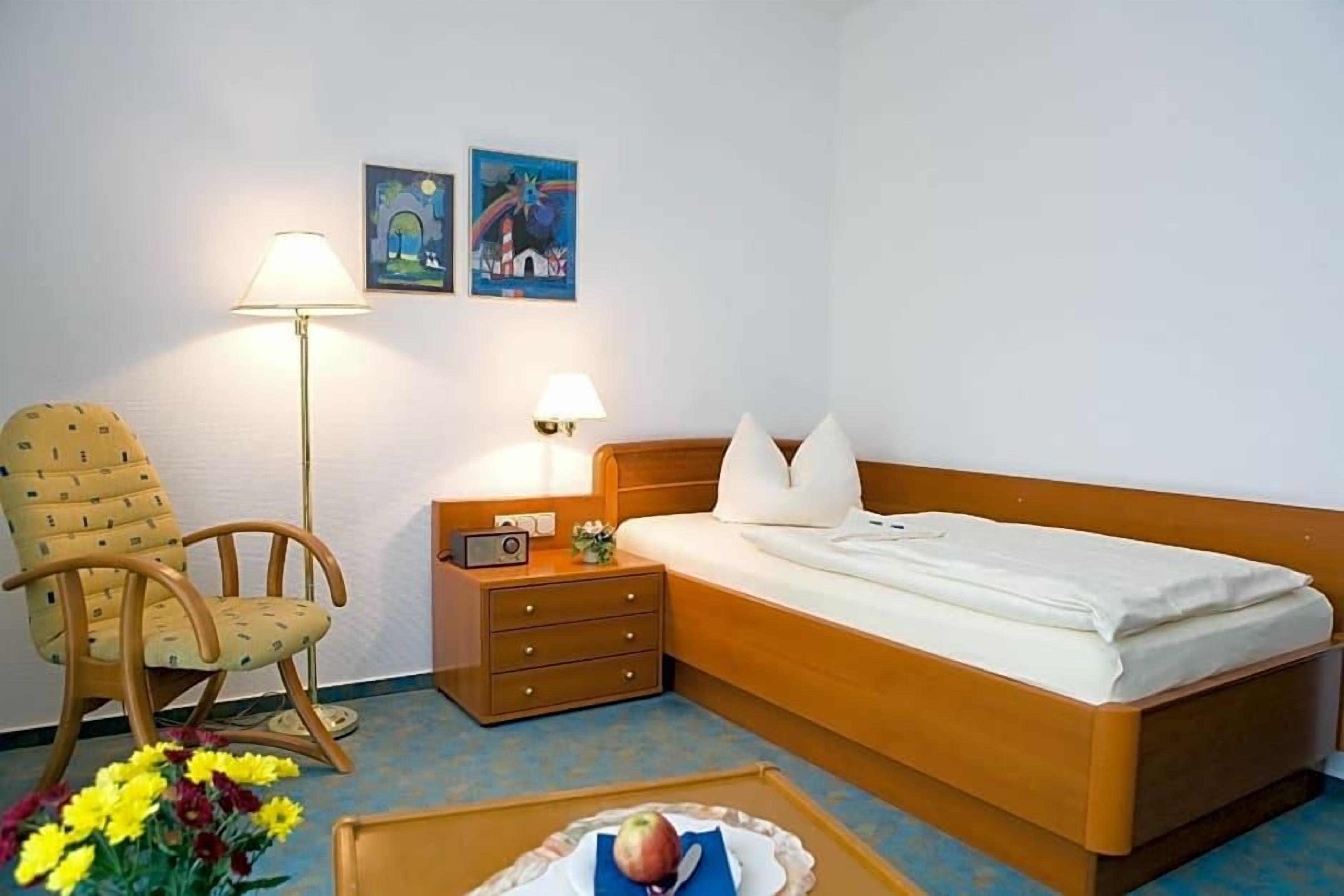 A hotel room with a single bed, a wooden nightstand, a lamp, a chair, and two wall art pieces.