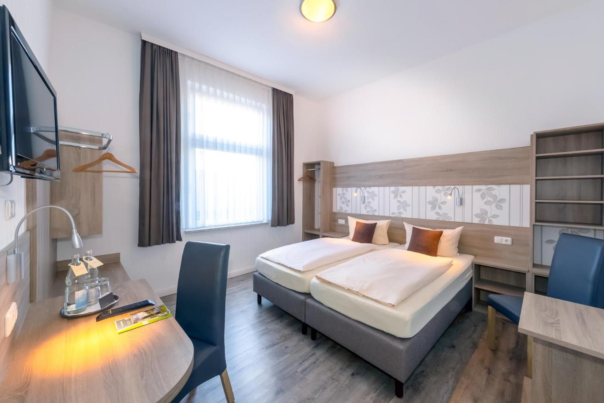 A hotel room with a bed, bedside tables, a desk, a chair, a wardrobe, and a window with curtains.