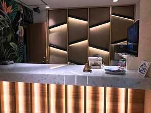 A hotel reception desk with a marble counter, a modern wall panel design, and a television screen.