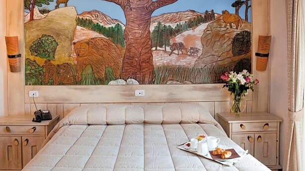 Une chambre à coucher ornée d’une grande fresque représentant une scène de forêt, un lit avec un plateau de petit-déjeuner, et des tables de chevet avec des lampes.