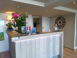 A reception desk with a clock, a potted plant, and informational brochures. - Hotel Le Lv Man (Saint-Gingolph)