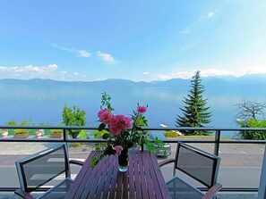 A balcony with a wooden table and a vase of flowers, overlooking a body of water and distant mountains. - Hotel Le Lv Man (Saint-Gingolph)