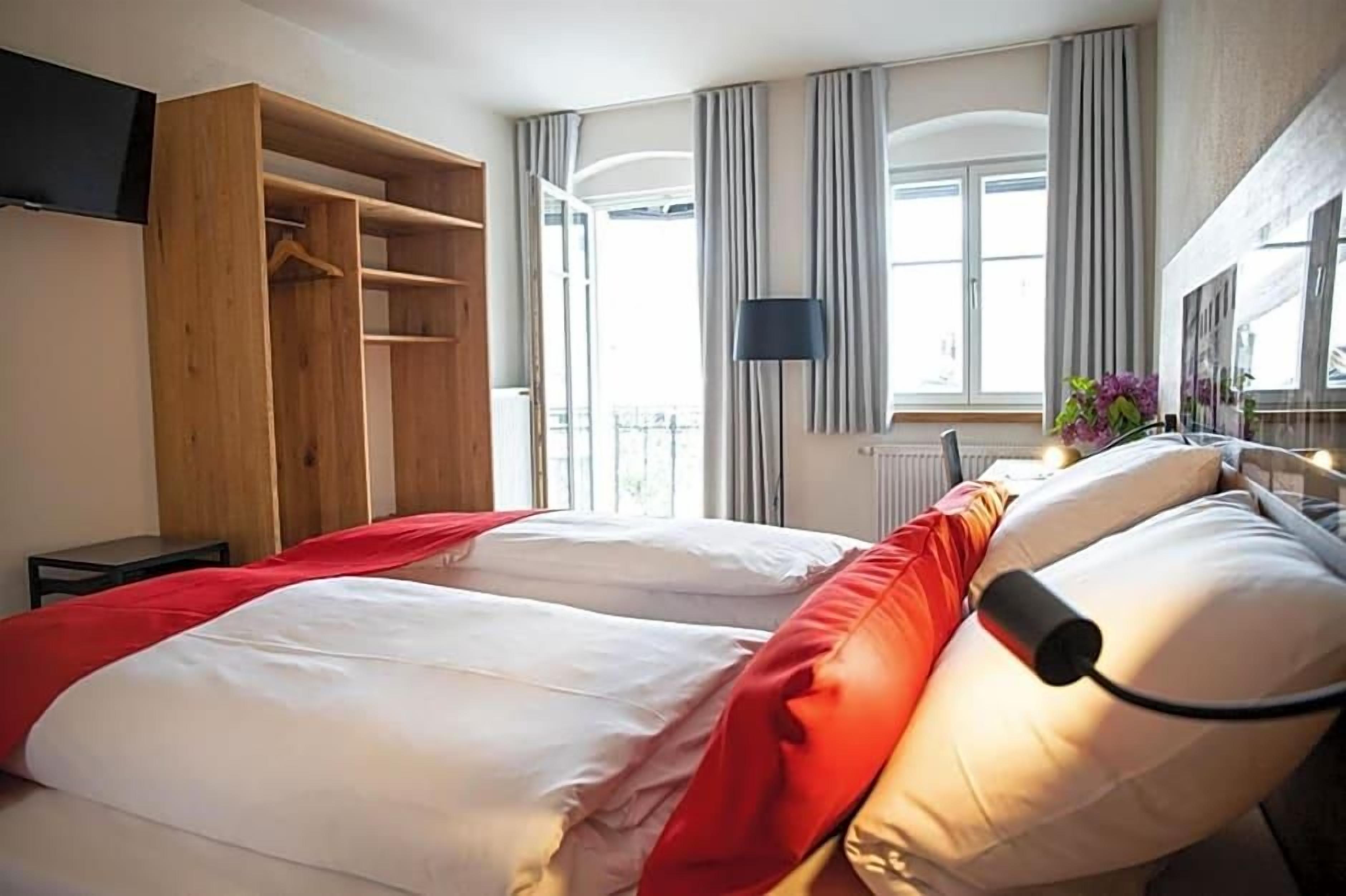 A hotel room with a bed, a wooden wardrobe, a window with curtains, and a bedside table.