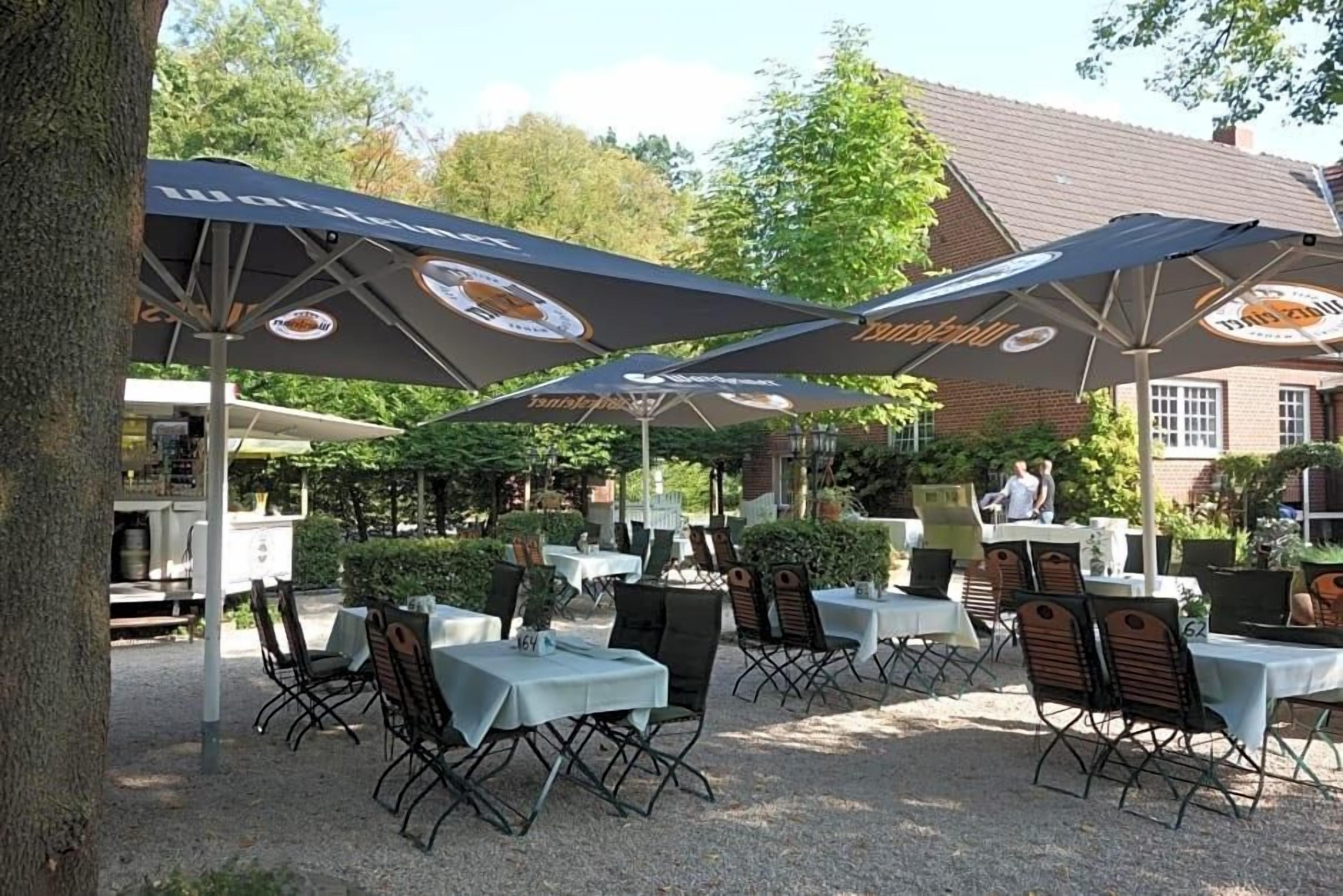 Ein Sitzbereich im Freien mit Tischen und Stühlen unter Sonnenschirmen, ein Gebäude im Hintergrund und ein Baum zur Linke.