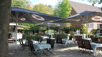 An outdoor seating area with tables and chairs under umbrellas, a building in the background, and a tree to the left.