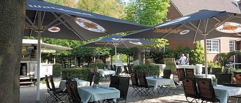 An outdoor seating area with tables and chairs under umbrellas, a building in the background, and a tree to the left.