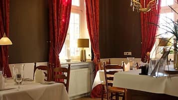 A dining area with tables set for two, red curtains, a chandelier, and a window with natural light.