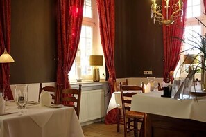 Comedor con mesas dispuestas para dos, cortinas rojas, candelabro y ventana con luz natural.