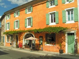 Un hotel con fachada tradicional, persianas verdes, mobiliario exterior y una zona de bar.