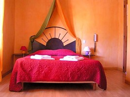 Una cama con una colcha roja, un cabecero con un diseño que recuerda al sol y una cortina con drapeado.