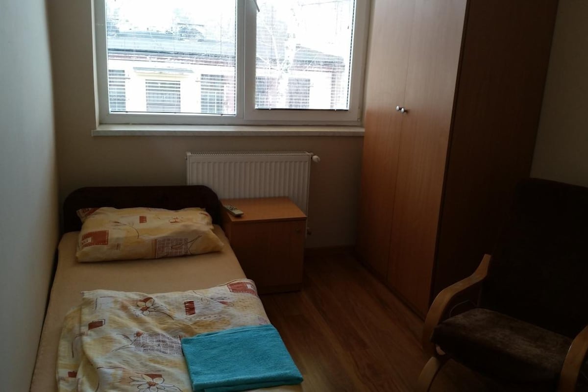 A small, single-bed room with a wooden floor, a window with blinds, a radiator, and a chair.