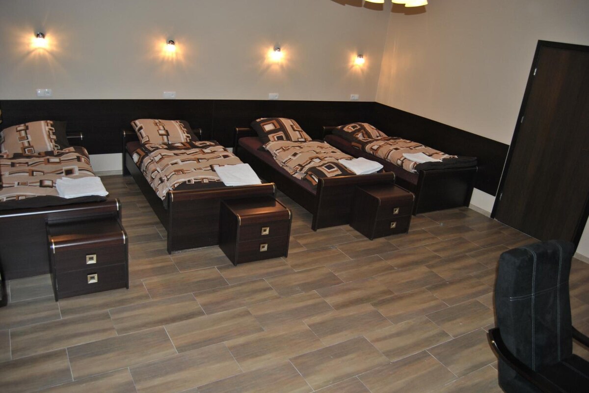 A room with four beds arranged in a semi-circle, each with a matching headboard and footboard. There are bedside tables with drawers, and the room has a wooden floor.