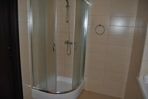 A glass-enclosed shower stall with a curved design, a white bathtub, and beige tiled walls. - Hotel Go Ociniec (Wloclawek)
