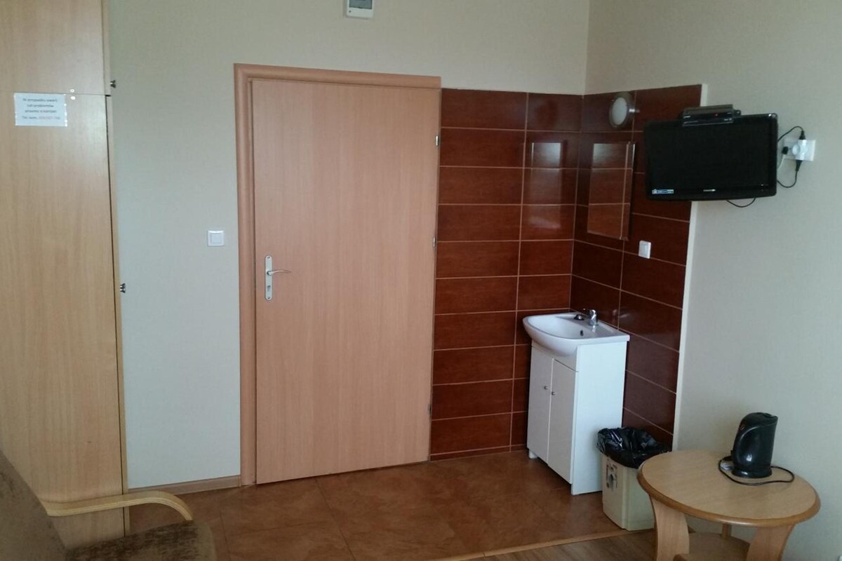 A room with a wooden door, a white sink, a small table with a black object, and a wall-mounted television.