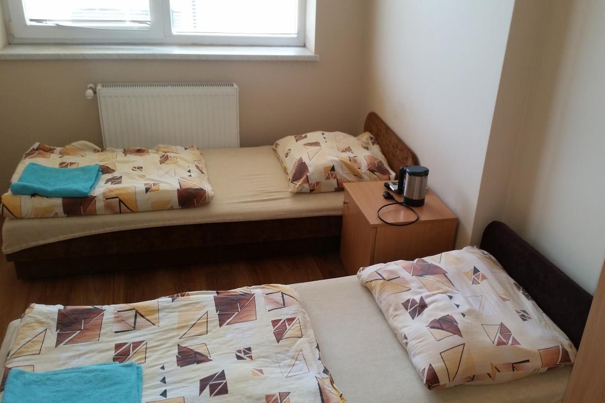 A small, compact room with two beds, a bedside table with a kettle, and a radiator.