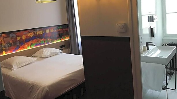 Una habitación de hotel con una cama, una lámpara de noche, una luz en la pared, un baño con lavamanos y una ventana.