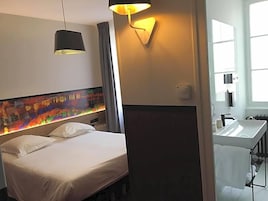 Una habitación de hotel con una cama, una lámpara de noche, una luz en la pared, un baño con lavamanos y una ventana.