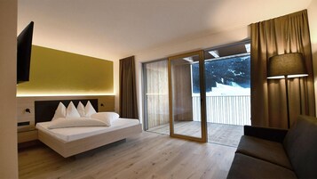 Quarto moderno com cama grande, mesa de cabeceira, televisão e varanda com vista para árvores.