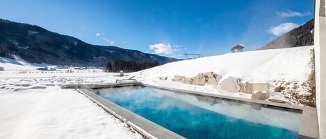 A snow-covered landscape with a clear blue pool, a stone structure, and a mountain backdrop.