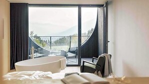 A modern bedroom with a large window, a bathtub, and a view of a balcony with outdoor furniture. - Hotel Milla Montis (Rio di Pusteria)