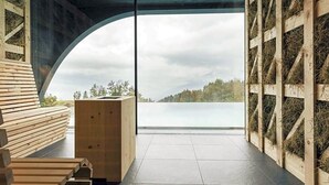 A modern interior with a large arched window overlooking a lake, wooden benches, and a central wooden structure. - Hotel Milla Montis (Rio di Pusteria)
