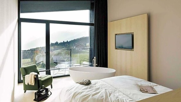 A modern bedroom with a large bed, a bathtub, a chair, and a balcony with a view. - Hotel Milla Montis (Rio di Pusteria)
