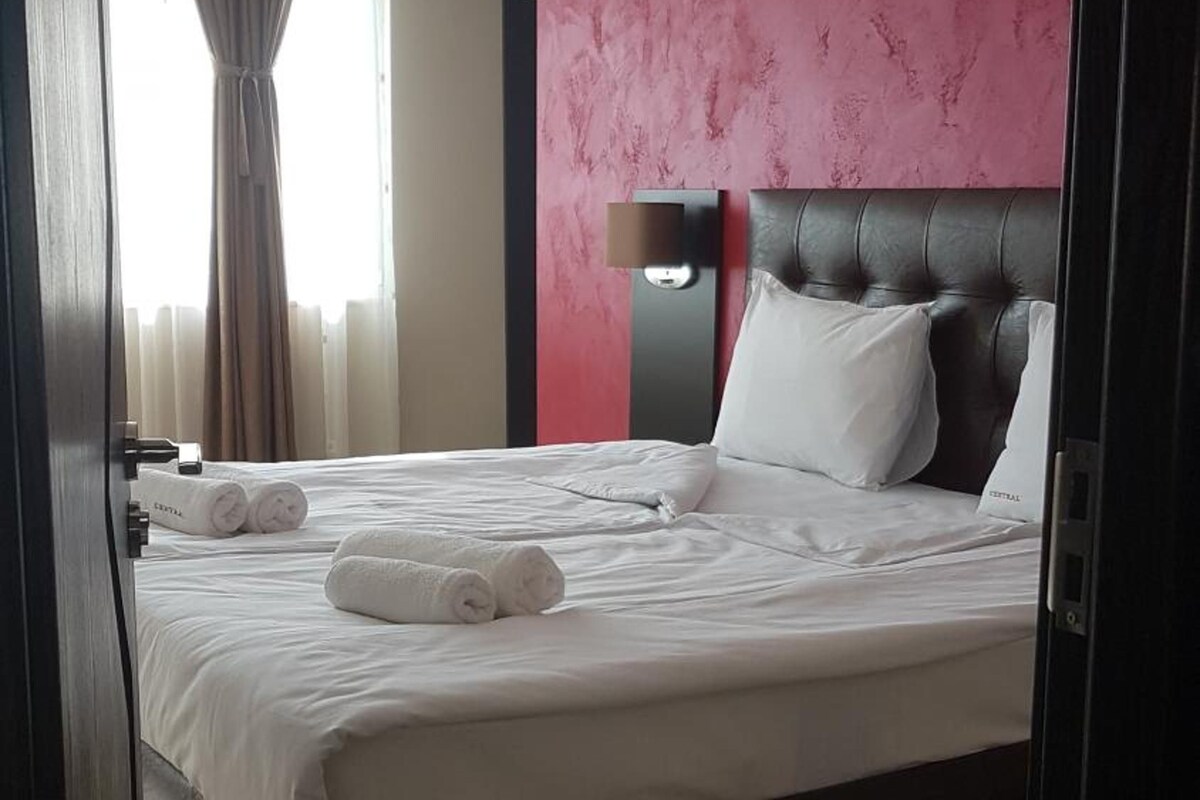 A hotel room with a large bed, a headboard, a nightstand, and a window with curtains.