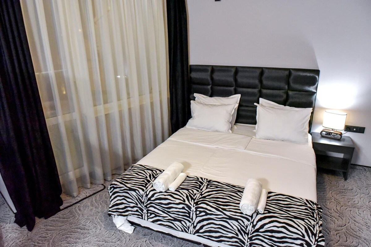 A hotel room with a bed, a nightstand, a lamp, and a window with curtains.