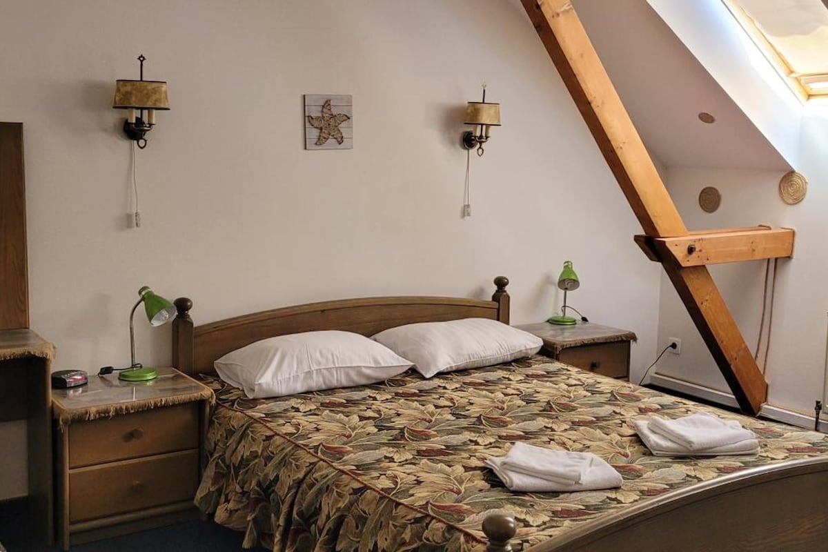 A bedroom with a sloped ceiling, a bed with a leaf-patterned bedspread, two wall-mounted lamps, a wooden chair, and a dresser.