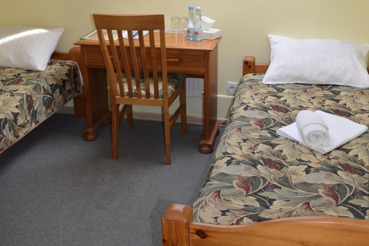 A small, single-room accommodation with a sloped ceiling and a skylight. There is a wooden desk with a chair, a bed with a floral bedspread, and a small table with a chair.