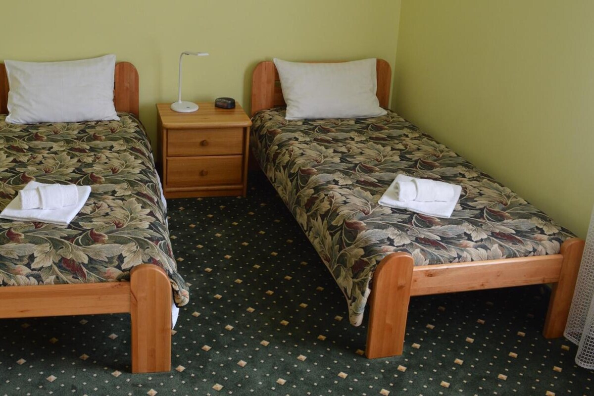 Una habitación con dos camas de madera, cada una con colcha estampada y almohada blanca. Entre las camas hay una mesita de noche de madera con una lámpara y un radio.