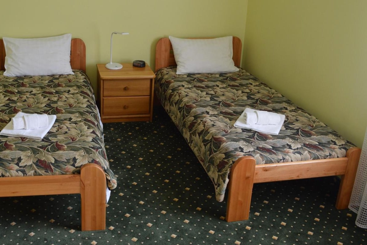 A room with two wooden beds, each with a floral bedspread and a white pillow. There is a small wooden nightstand with a lamp and a radio between the beds.