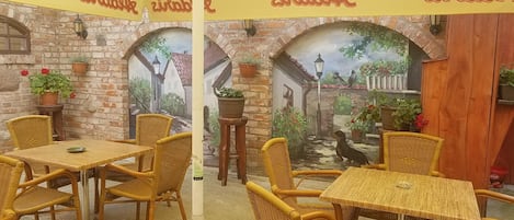 Un restaurante con terraza, mobiliario de mimbre y un mural que representa una escena de pueblo.