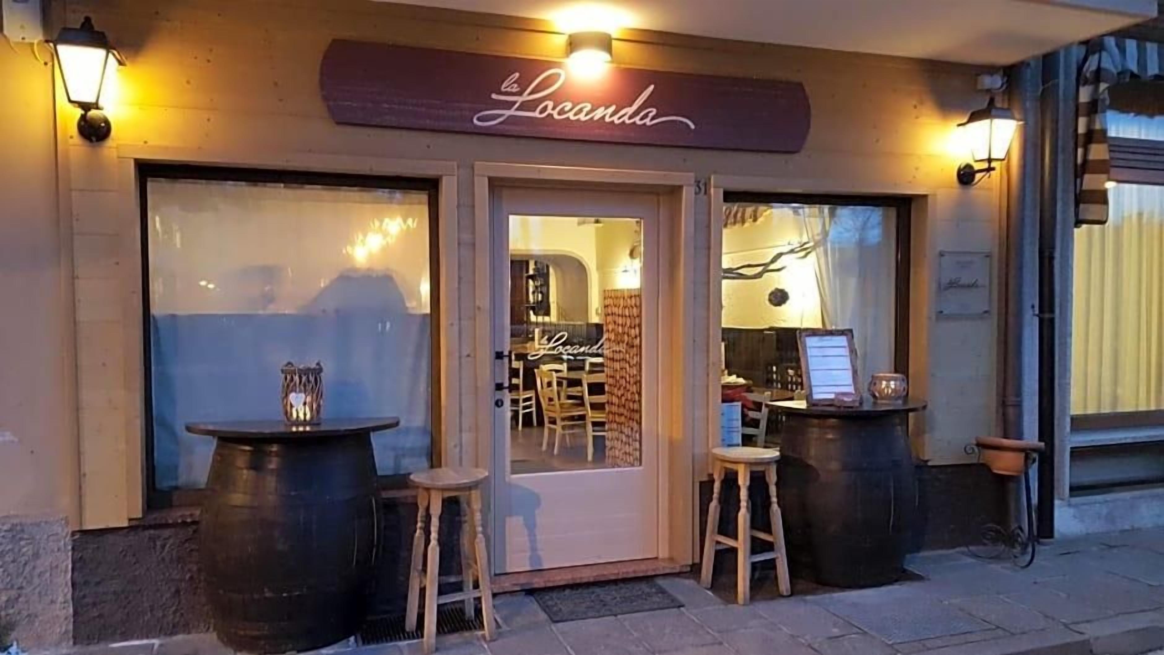 Um restaurante chamado 'La Locanda' com uma placa de madeira, dois bancos e uma mesa ao ar livre.